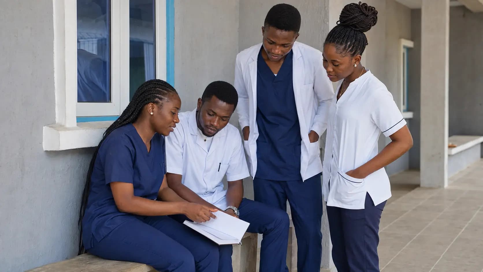 Students in navy uniforms and white clinical coats standing and talking together outside a classroom.