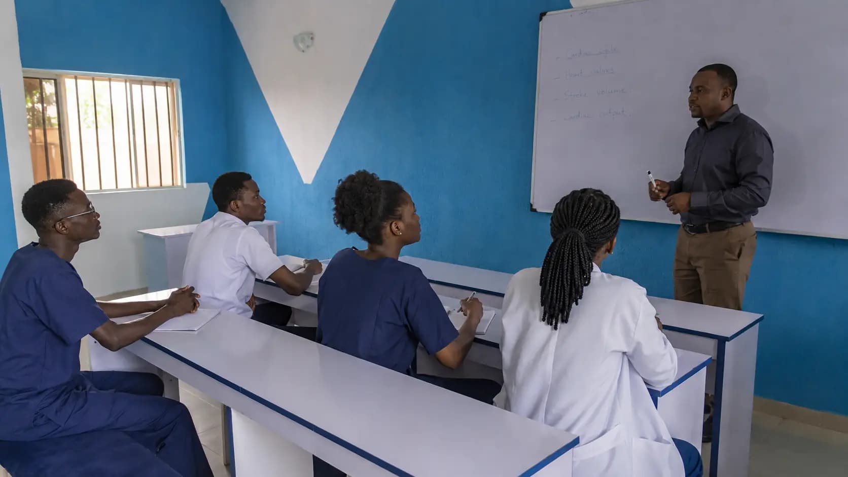 Students in navy scrubs and white uniforms seated with notebooks during a theory class in a classroom.